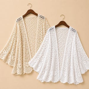May include: Two open-front cardigans, one in beige and one in white, featuring a delicate crochet knit pattern with wide, flared sleeves and a scalloped hemline. Each garment is displayed on a wooden hanger, showcasing the intricate floral-inspired lace texture and lightweight construction of the fabric.