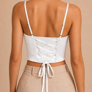 Op de afbeelding: Witte corset top met verstelbare spaghettibandjes en een veterrug. De top heeft een satijnen afwerking en een striksluiting in de taille. Het model draagt een beige broek met hoge taille.