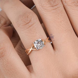 May include: A rose gold ring featuring a single round brilliant-cut white gemstone set in a four-prong setting. The band has a distinctive crisscross design that tapers towards the centre stone, creating a delicate and structured appearance.