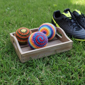 May include: Three colorful, crocheted balls in a wooden tray on green grass. The balls feature spiral patterns in various colors, including red, blue, yellow, and orange. Black and yellow soccer cleats are in the background.