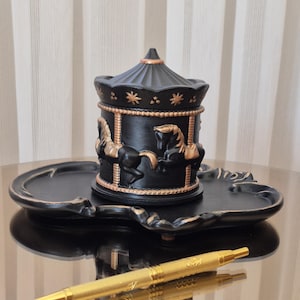 May include: A black carousel-style trinket box with gold detailing, decorated with horse motifs, is placed on a matching tray. A gold pen lies on the reflective surface, possibly a desk. The box has a domed lid.