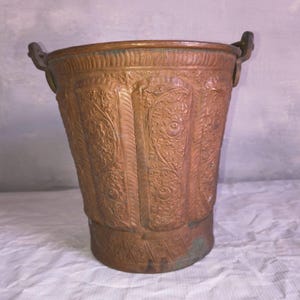 Può includere: Secchio color rame con motivi floreali e a foglie in rilievo. Il secchio ha due piccole maniglie e una forma leggermente svasata. Il metallo ha un aspetto invecchiato, che suggerisce l'età. Il design è simmetrico e dettagliato.