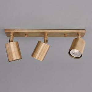 May include: A brass-colored, three-light track lighting fixture. The fixture has three adjustable cylindrical spotlights attached to a rectangular base. The lights are angled downward, and the fixture is mounted on a gray surface.