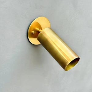 May include: A brass-colored wall-mounted spotlight fixture. The fixture has a circular base and a cylindrical light housing. The light housing is angled and has an open end. The fixture is mounted on a gray wall.