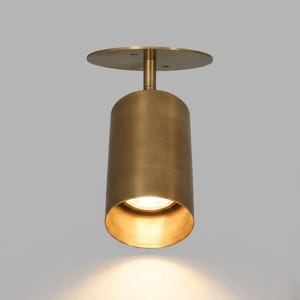 Op de afbeelding: Een messingkleurige, cilindrische plafondlamp met een cirkelvormige basis en een naar beneden gerichte lichtbron. De lamp heeft een eenvoudig, modern ontwerp en is geschikt voor diverse interieurs.