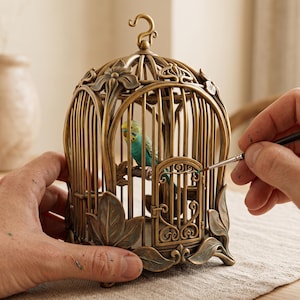 Pode incluir: Uma gaiola decorativa de metal em tom de bronze com motivos florais e de folhas detalhados na base e no topo. Dentro da gaiola, uma pequena figura realista de um periquito azul e amarelo está pousada num ramo.