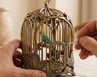 Puede incluir: Una jaula decorativa de metal en tono bronce con motivos florales y de hojas en la base y la parte superior. Dentro de la jaula, una pequeña figura realista de un periquito azul y amarillo se encuentra posada sobre una rama.