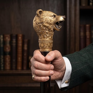 May include: A decorative walking stick with a gold-coloured bear head. The bear's head is highly detailed with an open mouth. The cane is held by a person wearing a green blazer and white shirt.