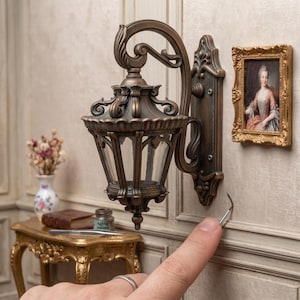 Op de afbeelding: Een miniatuur wandlantaarn met een donkerbronzen afwerking, voorzien van een sierlijke gebogen arm en een glazen kap met meerdere facetten. Het armatuur is gemonteerd op een rechthoekige achterplaat met decoratieve kruldetails en een punt.