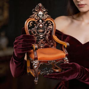 May include: Miniature ornate wooden chair with a skull on the backrest. The chair features intricate carvings, an orange seat, and a dark brown finish. The chair is held by gloved hands against a dark background.