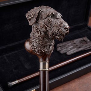 May include: A dark brown wooden walking stick featuring a detailed carved dog head handle. The handle includes an ornate collar design with a small shield emblem. The shaft is smooth, dark wood with a metallic band connecting to the handle.