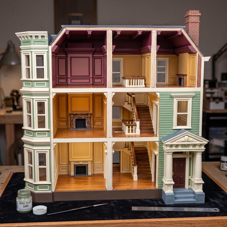 May include: A three-story dollhouse with a sage green exterior, white trim, and a brown front door. The interior features multiple rooms with wood-look flooring, fireplaces, and staircases. A small bottle of sage paint and a ruler are visible.