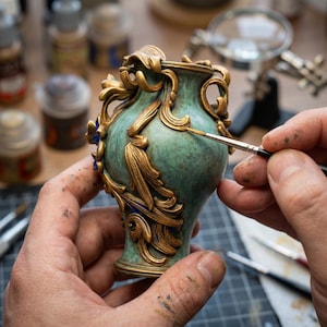 May include: A small, ornate vase with a teal base and gold filigree detailing. The vase is being hand-painted with a fine brush, highlighting the intricate design. The vase is approximately 4 inches tall.