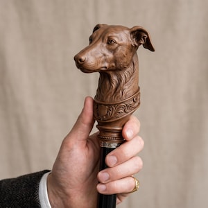 Op de afbeelding: Een bruine houten wandelstokgreep uitgesneden in de gedetailleerde vorm van een hondenkop. De basis van het snijwerk is voorzien van een sierlijk bloemmotief en een klein schildembleem, rustend op een gladde zwarte schacht met een zilverkleurige metalen ring.