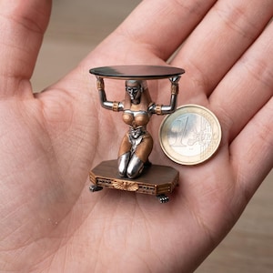 Puede incluir: Escultura en miniatura de una mujer arrodillada sosteniendo una pequeña mesa redonda. La figura está detallada con tonos plateados y dorados, arrodillada sobre una base rectangular. Una moneda de un euro está junto a la escultura.