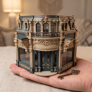 Op de afbeelding: Een miniatuur architectonisch model van een gebouw met blauwe en beige muren, gedetailleerde gouden accenten, boogramen en een donker dak. Een kleine, antiek ogende metalen sleutel met een ingewikkelde kop ligt naast de structuur.