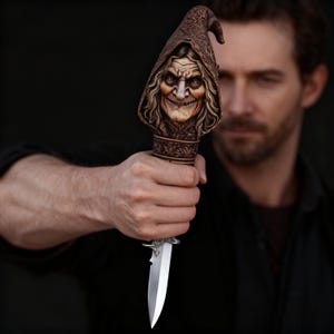 May include: A decorative dagger with a detailed handle featuring a carved witch's face under a pointed hood. The blade is silver, and the handle is brown with intricate carvings. The dagger is held against a dark background.