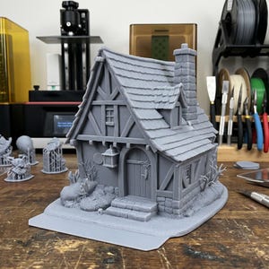 Può includere: Modello grigio stampato in 3D di un cottage stravagante. Il cottage presenta un tetto a falde ripide, un camino ed elementi architettonici dettagliati. Il modello include una porta d'ingresso, finestre e paesaggistica decorativa.