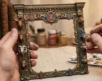 Puede incluir: Un marco de fotos rectangular con un acabado ornamentado en color bronce oscuro. El marco presenta cabezas de animales esculpidas que incluyen un alce, un ciervo, un carnero y lobos, junto con pequeños escudos heráldicos y un blasón central.