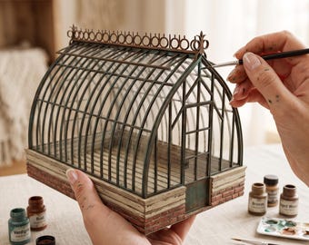 May include: A miniature greenhouse model with a dark metal frame, glass panels, and a brick-textured base. The structure features an arched roof with decorative metal trim along the top ridge and a small rectangular door on the front facade.