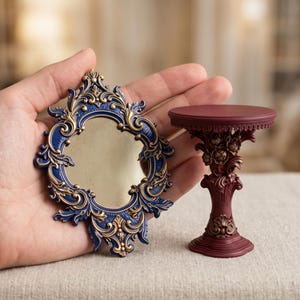 May include: A miniature oval mirror with a dark blue and gold ornate frame alongside a small, pedestal-style display stand in a deep maroon color with intricate floral and scrollwork detailing on the base and column.