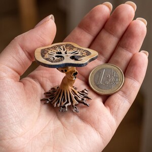 Puede incluir: Una escultura de madera en miniatura de un hongo con intrincados detalles tallados. El sombrero del hongo es de color marrón claro con un patrón oscuro, sostenido por un tallo y una base tallados. Una moneda de euro está al lado para comparar el tamaño.