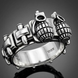 Può includere: Un anello d'argento con un design dettagliato di granate a mano in miniatura e forme geometriche. L'anello ha una finitura lucida ed è posto su uno sfondo scuro, evidenziando i suoi intricati dettagli.