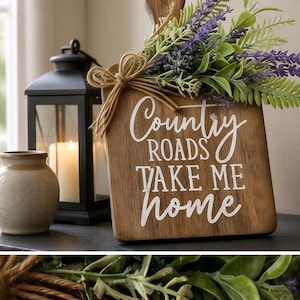 May include: A wooden cutting board with a handle, featuring the white text "Country ROADS TAKE ME home". The board is decorated with a jute twine bow and a cluster of artificial green leaves and purple lavender flowers attached to the top.
