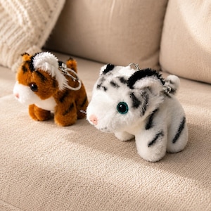 May include: Two plush tiger keychains, one with orange and black fur and the other with white and black fur. Both feature small ears, dark eyes, a pink nose, and a metal keyring attached to the top of their heads, showcasing a soft texture.