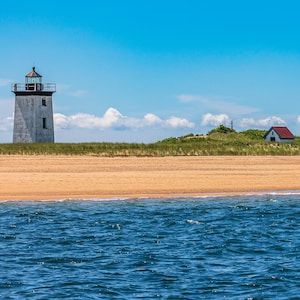 Könnte beinhalten: Eine Küstenlandschaft mit einem weißen Leuchtturm und einem kleinen Gebäude mit rotem Dach an einem Sandstrand. Der Vordergrund zeigt blaues Meerwasser unter einem klaren, hellblauen Himmel mit verstreuten Wolken. Der Leuchtturm steht links.