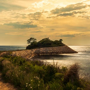 Könnte beinhalten: Eine Küstenlandschaft mit einem Feldweg, der zu einer kleinen, erhöhten Landmasse führt, die mit Steinmauerwerk befestigt ist. Das Gebiet ist mit grüner Vegetation und Bäumen bewachsen und vom Ozean unter einem goldenen Sonnenuntergangshimmel umgeben.