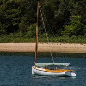 Op de afbeelding: Een kleine zeilboot met een witte romp en houten accenten drijft op blauw water. De boot heeft een hoge houten mast en een groene canvas afdekking. Een zandstrand en groene bomen zijn op de achtergrond.