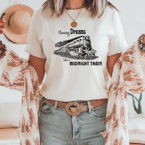 May include: Cream-colored t-shirt featuring a black and white illustration of a vintage train. The text "Chasing Dreams Like a Midnight Train" is printed above and below the train graphic. The shirt is paired with blue jeans and a brown belt.