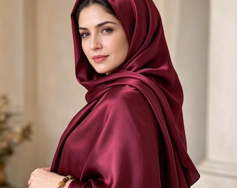 Könnte beinhalten: Ein tiefburgunderrotes Satin-Kopftuch, das über Kopf und Schultern drapiert ist und eine glatte, reflektierende Stofftextur aufweist. Das Tuch wird mit einem Perlenarmband aus runden, erdfarbenen Steinen am Handgelenk getragen.