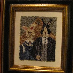 May include: A framed needlepoint artwork depicting two rabbits dressed in formal attire. The rabbit on the left is wearing a blue dress with white trim, while the rabbit on the right is wearing a black suit with a white shirt and bow tie. The artwork is set against a neutral background.