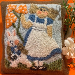 May include: A white and blue needlepoint pillow featuring Alice in Wonderland with a white rabbit and orange mushrooms. The pillow is framed with a brown border.