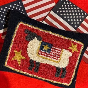 May include: A red, white and blue sheep-shaped cushion with gold stars. The sheep is wearing a small American flag on its back. The cushion is on a red background.