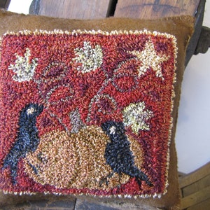 Puede incluir: Un cojín cuadrado marrón con un diseño cosido a mano. La imagen central presenta dos pájaros negros posados sobre una calabaza beige, rodeados de follaje rojo y acentos blancos en forma de estrella. El borde del cojín es marrón claro.