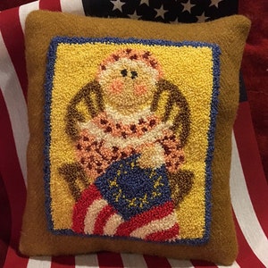 May include: A brown cushion with a yellow background and a blue frame. The cushion features a needlepoint design of a woman sitting in a rocking chair holding a flag with stars and stripes.