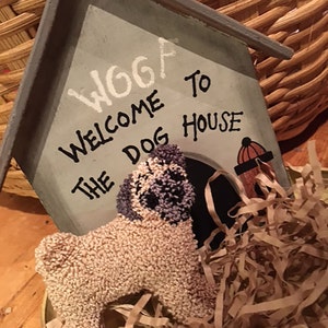 May include: A wooden dog house painted grey with the words "Woof Welcome to the Dog House" in black paint. A small beige plush pug dog is sitting in front of the dog house.