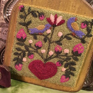 Puede incluir: Alfombra cuadrada cosida a mano con un diseño floral y de pájaros. La alfombra presenta un corazón central en rojo oscuro, rodeado de flores rosas, pájaros azules y follaje verde sobre un fondo verde claro. La alfombra está montada sobre una tabla de madera.