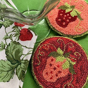 May include: Two round, crocheted coasters with a strawberry design. The coasters are pink and green with a red strawberry in the center.