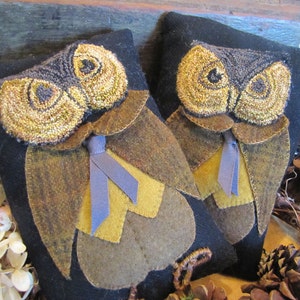 Puede incluir: Dos almohadas de fieltro con forma de búho con detalles en marrón, negro y dorado. Los búhos tienen ojos grandes y redondos y una cinta azul atada alrededor de sus cuellos.