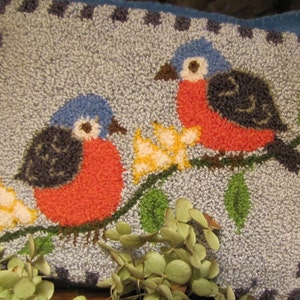 Puede incluir: Una almohada bordada en azul y rojo con dos pájaros posados en una rama con hojas verdes y flores amarillas.