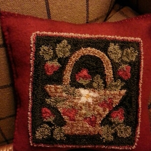 May include: A red felt pillow with a hand-stitched design of a basket filled with strawberries and green leaves. The basket is brown and has a white flower in the center.