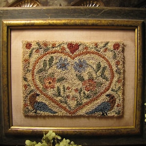 Puede incluir: Una obra de arte de punto de aguja enmarcada que presenta un diseño en forma de corazón con flores y pájaros. El corazón está delineado en marrón y relleno con una variedad de colores, incluidos el verde, el rojo y el azul. Los pájaros están posados ​​en ramas y tienen cuerpos azules y cabezas marrones.