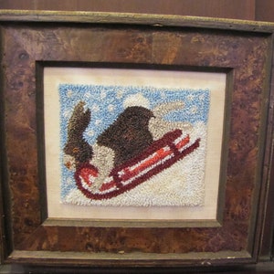 May include: A framed needlepoint picture of a brown bear wearing a white hat and riding a red sled down a snowy hill.