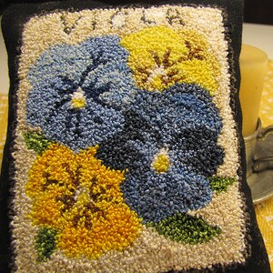May include: A close-up of a hand-stitched needlepoint cushion featuring three stylised pansies in shades of blue, yellow, and green. The cushion is framed with a white border and a black outer edge.