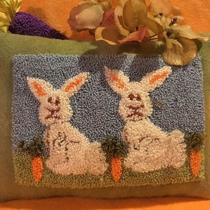 May include: A green pillow with a blue background and two white bunnies with orange ears. The bunnies are sitting in front of orange carrots in a green field.