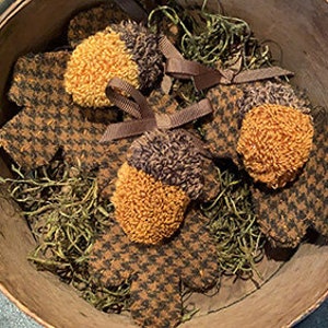 Puede incluir: Tres adornos de bellota de fieltro marrón y naranja con lazos de cinta marrón. Las bellotas están unidas a hojas de roble de fieltro marrón y blanco a cuadros.